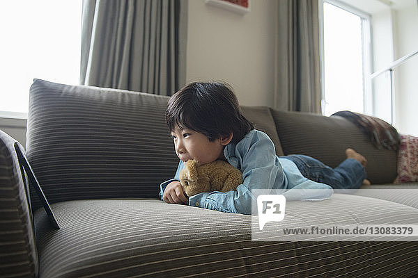 Junge sieht Tablette an  während er mit Stofftier auf dem Sofa zu Hause liegt