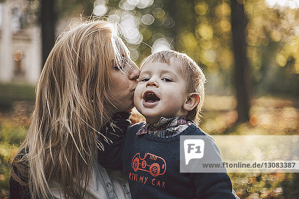 Porträt eines süßen Sohnes  während seine Mutter ihn im Herbst im Park küsst