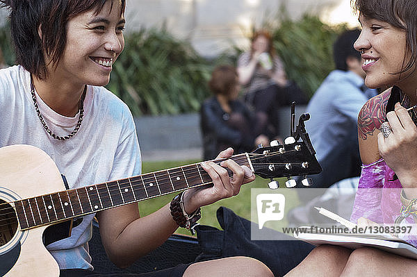 Lächelnde Frau hört einem Freund zu  der im Park Gitarre spielt
