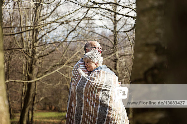 In eine Decke gewickeltes älteres Paar  das sich im Winter an kahlen Bäumen umarmt