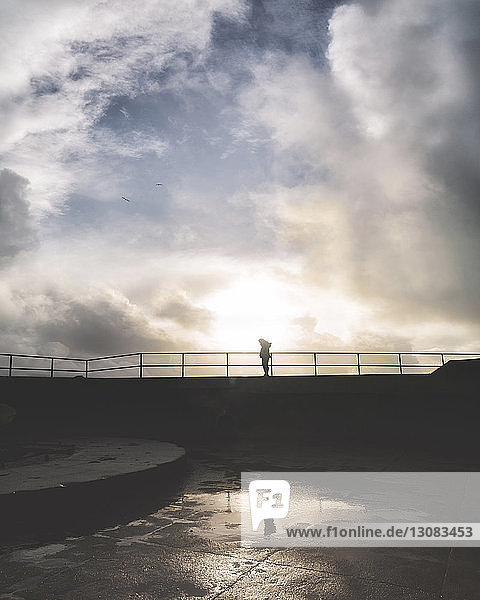 Silhouettenfrau steht bei Sonnenuntergang am Geländer vor bewölktem Himmel