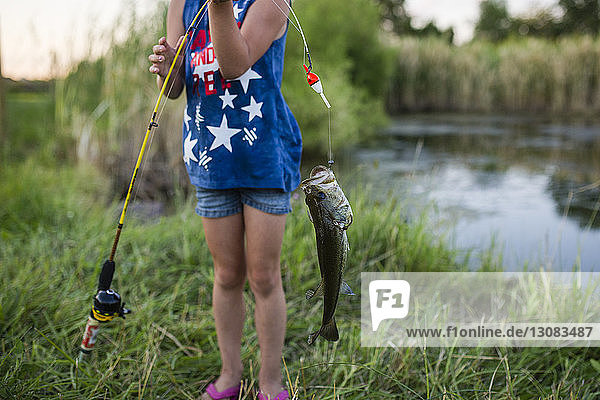 Mädchen  das am See stehend einen gefangenen Fisch mit einer Angelrute hält