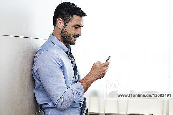 Seitenansicht eines Geschäftsmannes  der ein Smartphone benutzt  während er sich im Hotelzimmer an die Wand lehnt
