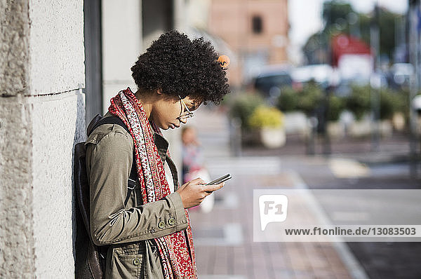 Side view of woman using phone while leaning on wall at sidewalk