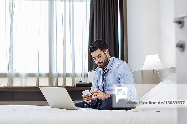 Geschäftsmann benutzt Smartphone  während er im Hotelzimmer auf dem Bett sitzt
