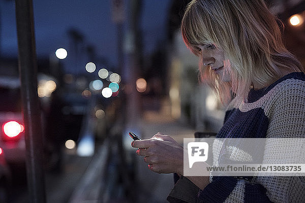 Seitenansicht einer Frau  die ein Smartphone in einer beleuchteten Stadt bei Nacht benutzt