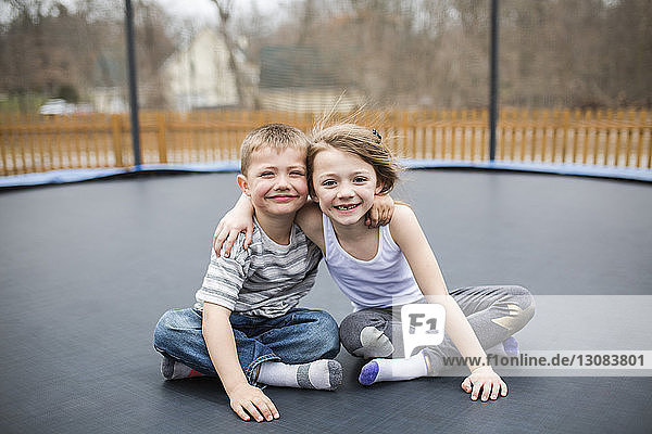 Porträt von glücklichen Geschwistern  die auf dem Spielplatz auf einem Trampolin sitzen