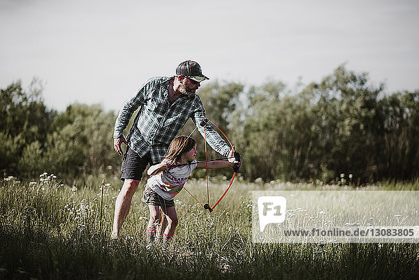 Father helping daughter in using bow and arrow at grassy field