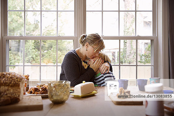 Liebende Mutter umarmt Sohn  während sie am Essenstisch am Fenster im Wohnzimmer sitzt