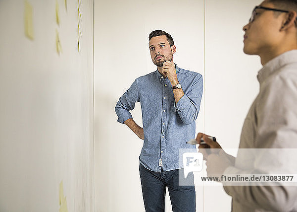 Business people discussing over sticky notes while standing against white wall in creative office