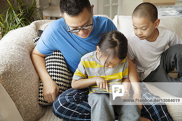 Father looking at daughter using tablet computer while sitting with son on sofa