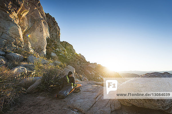 Hiker sitting on rock formation by mountains against clear sky on sunny day