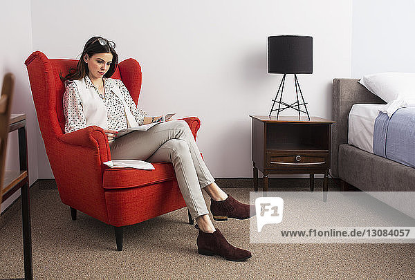 Businesswoman reading magazine while sitting on red armchair in hotel room