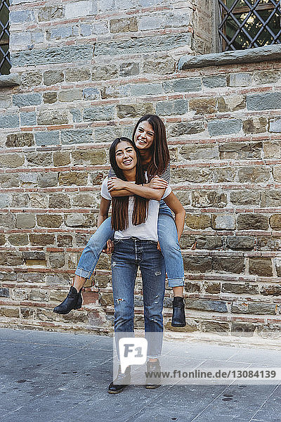 Portrait of happy woman piggybacking friend while standing on street by building