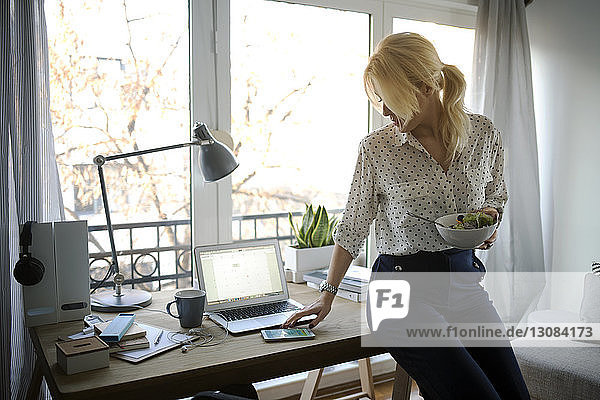 Frau benutzt Smartphone  während sie im Heimbüro eine Salatschüssel in der Hand hält