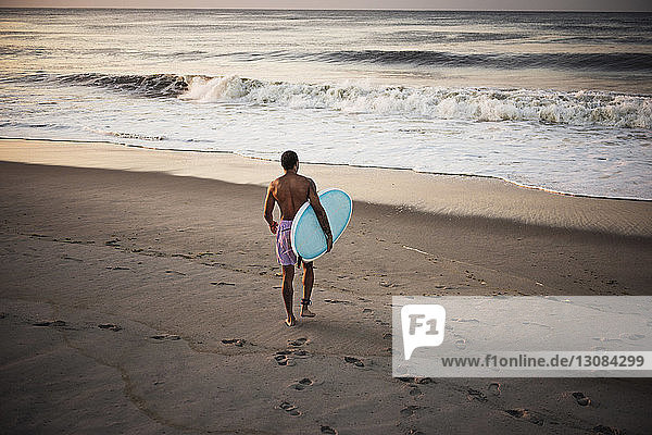 Rückansicht eines Mannes  der ein Surfbrett trägt  während er am Strand auf Sand läuft