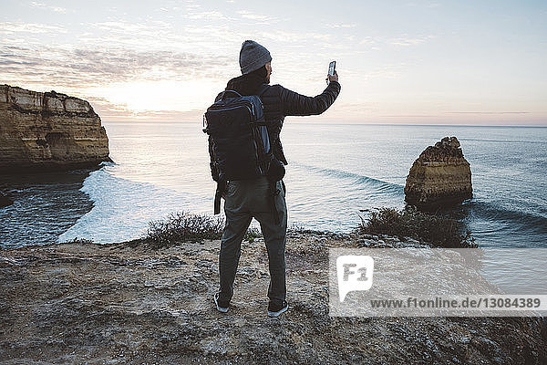 Rückansicht eines Mannes  der sich mit einem Smartphone selbstständig macht  während er bei Sonnenuntergang am Strand gegen den Himmel steht