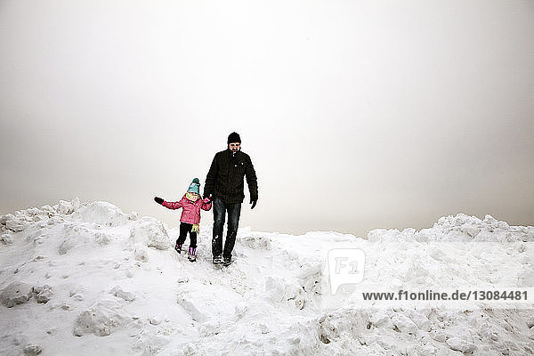 Grandfather and granddaughter walking on snow covered field against cloudy sky