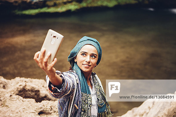 Schrägaufnahme einer Frau  die sich am Flussufer stehend per Mobiltelefon selbstständig macht