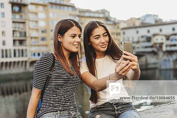 Freunde nehmen Selfie mit dem Handy  während sie auf einer Stützmauer am Fluss Arno in der Stadt sitzen