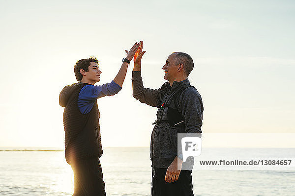 Glückliche Vater und Sohn machen bei Sonnenuntergang ein High-Five gegen Meer und Himmel