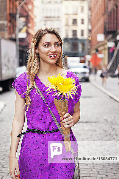 Lächelnde Frau hält Blumenstrauss beim Spaziergang auf gepflasterter Straße in der Stadt