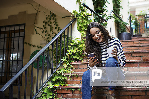 Lächelnde Frau benutzt Mobiltelefon und hält Kaffeetasse  während sie auf Stufen sitzt