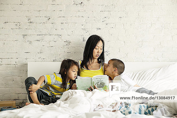Mutter liest Kindern ein Buch vor  während sie zu Hause im Bett an der Wand sitzt