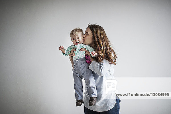 Mother kissing baby boy while standing against white wall at home