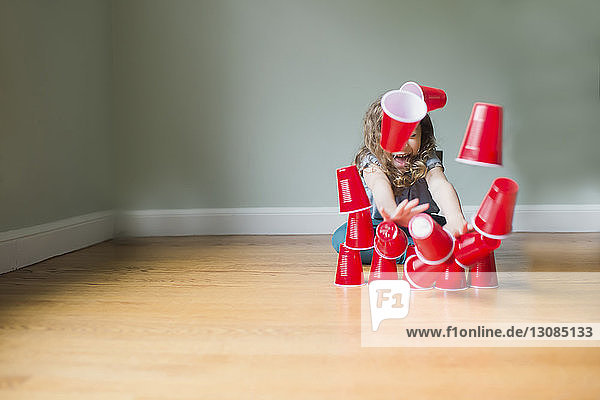 Playful girl breaking pyramid made of disposable cups while sitting on floor at home