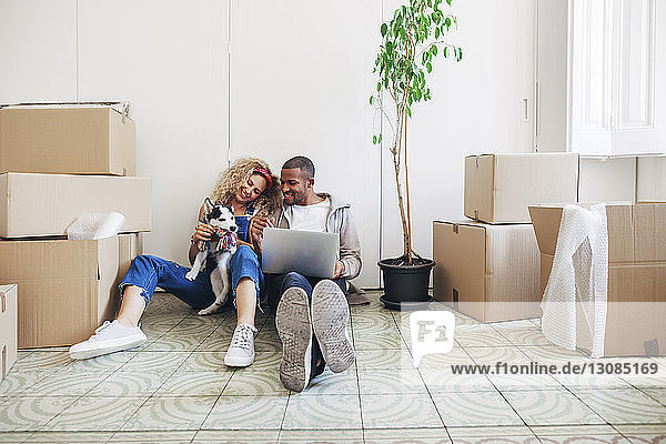 Happy couple playing with dog while using laptop computer at new home