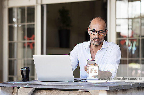 Geschäftsmann benutzt ein Smartphone  während er mit seinem Laptop am Tisch im Büro sitzt