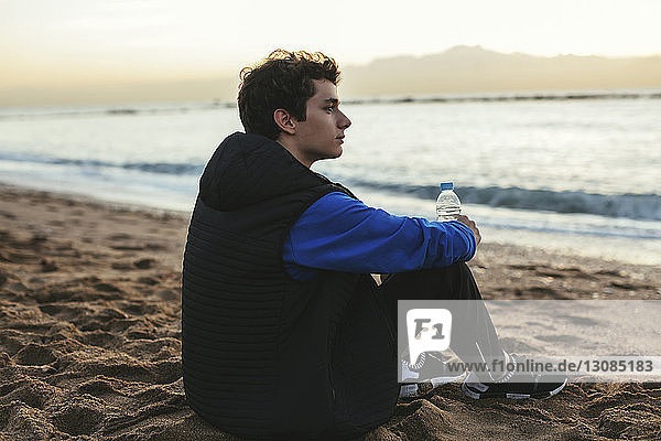 Side view of thoughtful teenage boy holding water bottle while sitting at beach