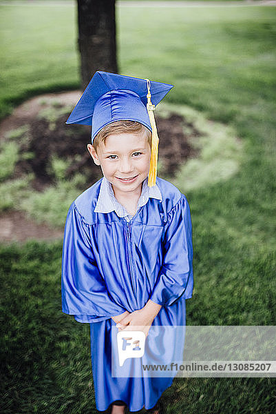 Hochwinkelporträt eines Jungen im Graduiertenkleid  der auf einem Grasfeld im Park steht