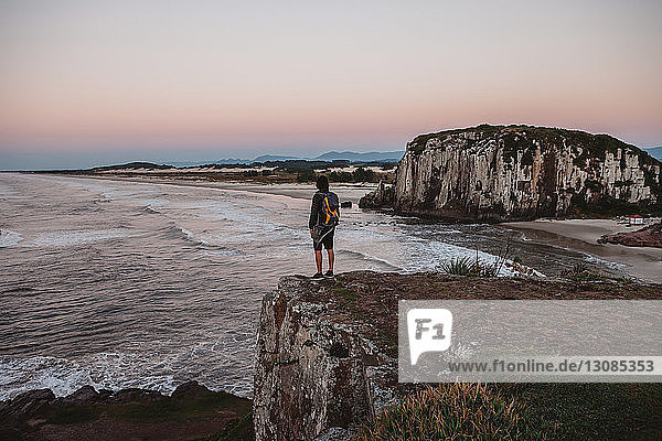 Rear view of male hiker standing on cliff at beach during sunset