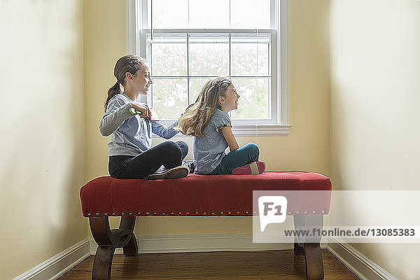 Side view of girl combing sister's hair while sitting on seat against window at home