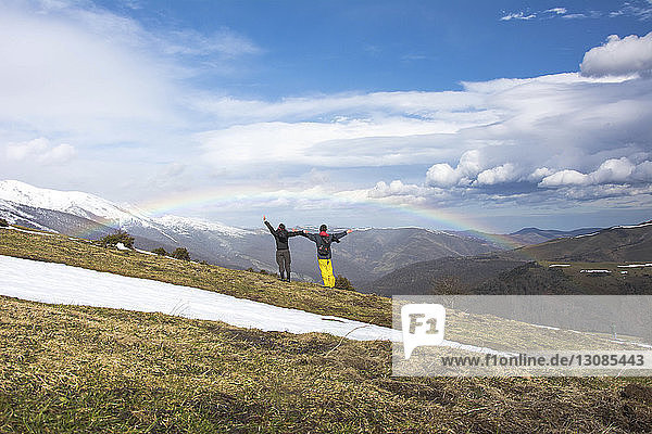Rear view of friends with arms outstretched standing against mountains and rainbow during winter