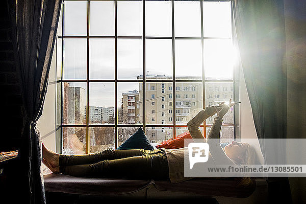 Frau benutzt Tablet-Computer  während sie zu Hause auf einem Fensterplatz in einer Nische liegt