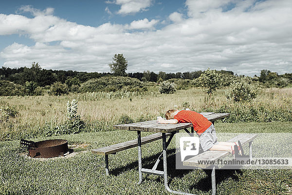 Side view of boy lying on picnic bench against cloudy sky during sunny day