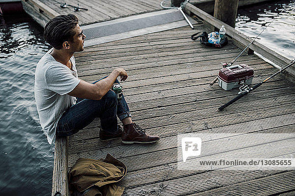 High angle view of thoughtful man holding drink can while sitting on pier over river