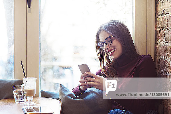 Glückliche Frau benutzt Mobiltelefon  während sie im Cafe am Fenster sitzt