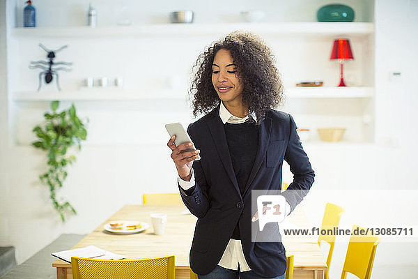 Lächelnde Geschäftsfrau benutzt Mobiltelefon  während sie im Büro am Tisch steht
