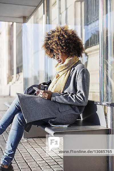 Frau benutzt Mobiltelefon  während sie auf dem Sitz an der Bushaltestelle sitzt