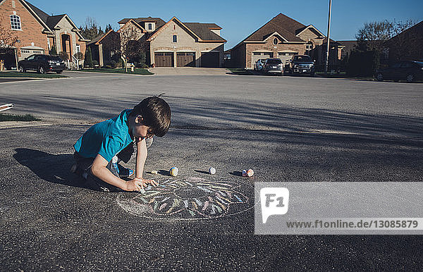 Side view of boy drawing with chalk on asphalt against houses