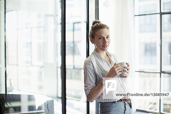 Porträt einer Geschäftsfrau  die etwas trinkt  während sie sich im Büro ans Fenster lehnt