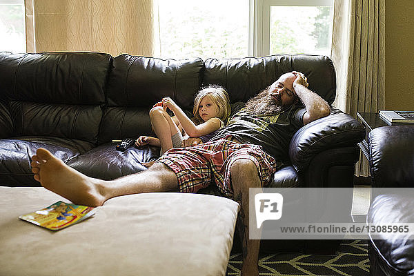 Mädchen schaut weg  während sie mit dem auf dem Sofa schlafenden Vater sitzt