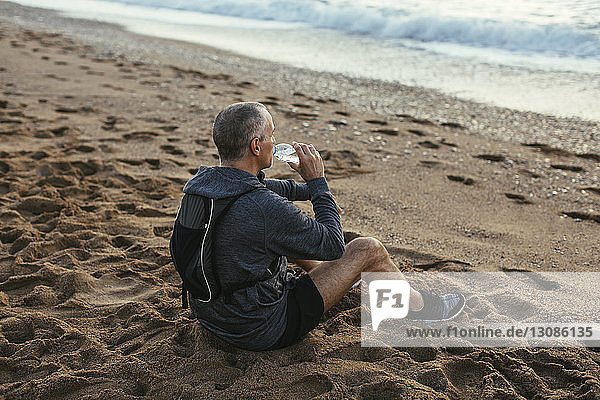 Hochwinkelaufnahme eines Mannes  der am Strand sitzt und Wasser trinkt