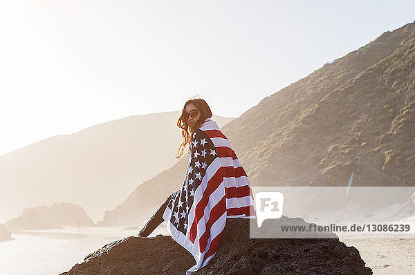 Porträt einer in eine amerikanische Flagge gehüllten Frau  die auf einem Felsen am Strand sitzt