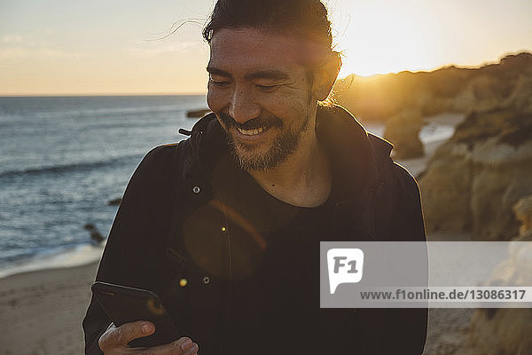 Lächelnder Mann benutzt Smartphone  während er bei Sonnenuntergang am Strand steht