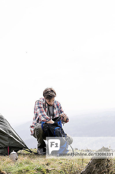 Mann sucht im Rucksack auf einem Campingplatz auf einem Hügel bei klarem Himmel